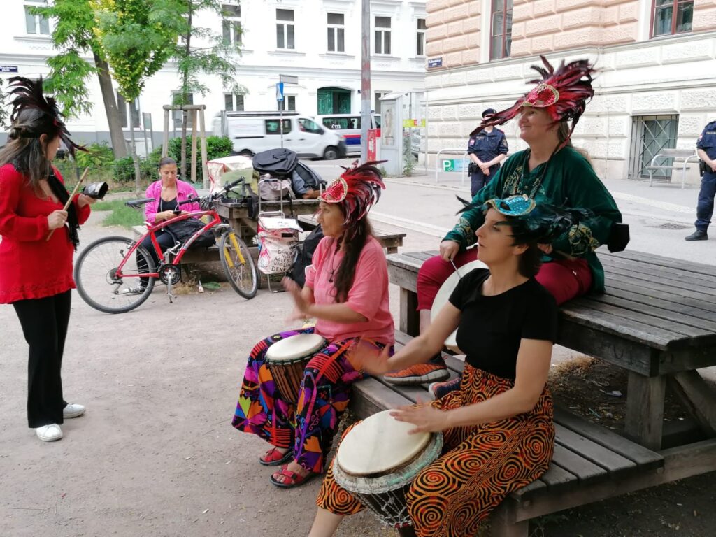 Die Rhythm Sistas Unlocked spielen auf der Bank eines Jausentisches vor einer Schule mit den Instrumenten Djembe, Rahmentrommel und Glocke.